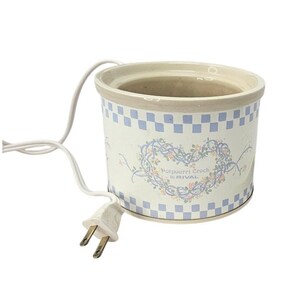 May include: A cylindrical ceramic potpourri crock by Rival with a white base, a light blue checkered border, and a central floral heart design. The crock features a white electrical cord with a two-prong plug extending from the side.