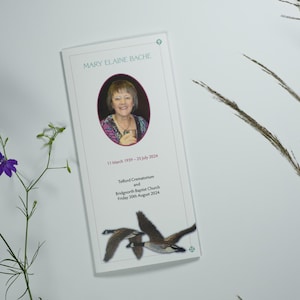 May include: A white memorial card featuring a portrait of Mary Elaine Bache, with dates and service details. The card is decorated with a pair of flying geese. Purple flowers and grasses are in the background.
