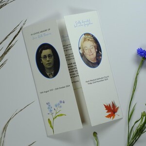 May include: Two memorial cards with oval portraits and text. One card has a photo of a person wearing glasses, the other has a photo of a person with glasses. The cards are decorated with floral and leaf illustrations. Blue flowers and grass are in the background.