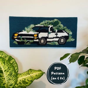 May include: Crocheted wall hanging featuring a white and black car with a green tree on top, set against a teal background. The car has yellow headlights and gray wheels. A circular graphic reads "PDF Pattern (en & fr)".