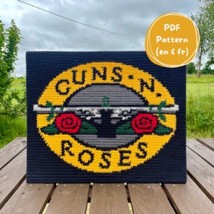May include: A navy blue crochet piece displaying the Guns N' Roses logo. The logo features a yellow circle with the band's name, a grey gun design, and red roses. The text "PDF Pattern (en & fr)" is in a yellow circle.