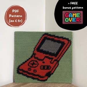 May include: A handmade crochet piece featuring a red video game console design on a green background. The image includes the text "PDF Pattern (en & fr)" and a bonus pattern with the words "GAME OVER".