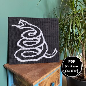 May include: A square crochet wall hanging featuring a coiled snake design in white yarn against a solid black background. A circular black badge in the corner contains the white text PDF Pattern (en & fr), indicating a digital download file.