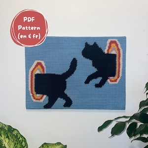 May include: A blue crocheted wall hanging featuring two black cat silhouettes stepping through colorful rainbow portals. The text "PDF Pattern (en & fr)" is in a red circle in the upper left corner. The artwork is displayed on a white wall.