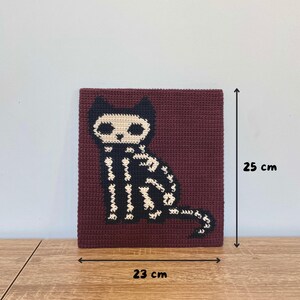Peut inclure: Carr&eacute; crochet&eacute; avec un motif de chat squelette noir et blanc sur fond bordeaux. L'&oelig;uvre mesure environ 23 cm de large et 25 cm de haut. Le chat est assis droit, la queue enroul&eacute;e.