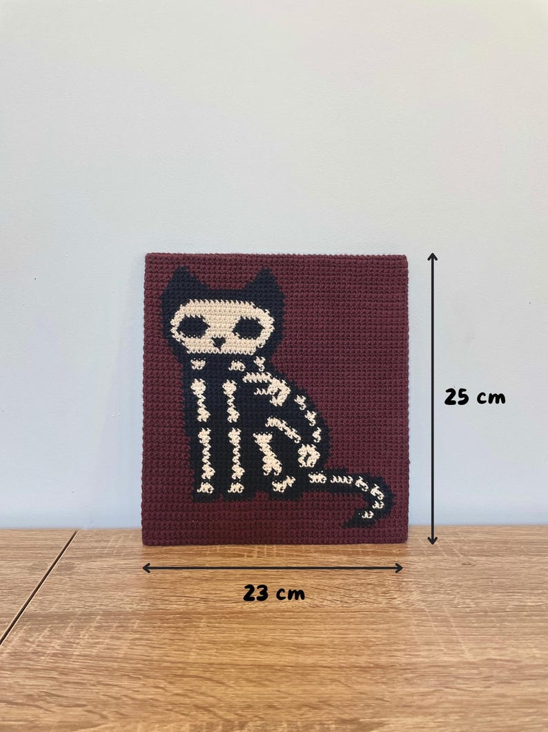 Peut inclure: Carr&eacute; crochet&eacute; avec un motif de chat squelette noir et blanc sur fond bordeaux. L'&oelig;uvre mesure environ 23 cm de large et 25 cm de haut. Le chat est assis droit, la queue enroul&eacute;e.