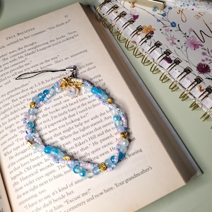 May include: A delicate beaded bracelet featuring light blue, white, and pink beads, accented with gold-colored heart charms. The bracelet is displayed on an open book with visible text and a floral-patterned notebook in the background.
