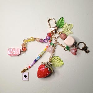 May include: A whimsical keychain featuring a red strawberry charm, a miniature milk carton, and a playing card with a spade. The keychain includes colorful beads, gold-tone hardware, and various decorative charms, including leaves and flowers.