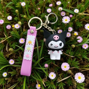 May include: A pink keychain with a black and white cartoon character charm. The keychain has a pink strap with white scalloped edges and decorative elements. The charm is a black character with a skull-like design and a white body. The background is green grass and small flowers.