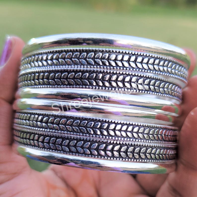 May include: A wide silver cuff bracelet with a detailed leaf pattern. The bracelet has a polished, shiny finish and is made of multiple bands. The leaf pattern is repeated around the bracelet.