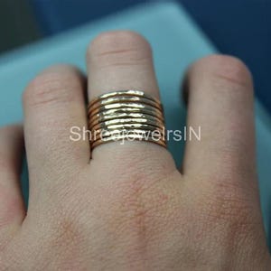 May include: A stackable gold ring with a hammered texture. The ring is composed of multiple bands, creating a wide, textured appearance. The ring is worn on a finger, showcasing its design and craftsmanship. The ring is a warm gold color.
