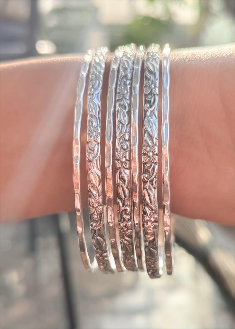 Peut inclure: Plusieurs bracelets en argent, chacun orn&eacute; d'un motif floral d&eacute;taill&eacute;. Les bracelets sont empil&eacute;s sur un poignet, mettant en valeur leur design complexe et leur surface r&eacute;fl&eacute;chissante. La lumi&egrave;re met en &eacute;vidence les fleurs et les feuilles en relief.