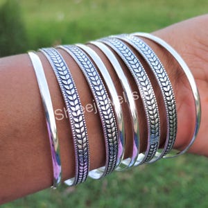 May include: A collection of silver bangles. Some bangles are plain, while others feature a detailed leaf pattern. The bangles are worn on a wrist, with a blurred green background.