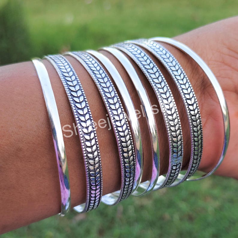 May include: A collection of silver bangles. Some bangles are plain, while others feature a detailed leaf pattern. The bangles are worn on a wrist, with a blurred green background.