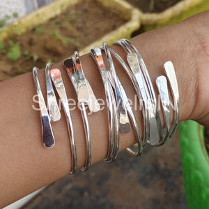 Può includere: Una collezione di braccialetti in metallo color argento con una texture martellata. I braccialetti sono indossati su un polso, mostrando il loro design a polsino aperto e le superfici riflettenti. I gioielli sono semplici ed eleganti.