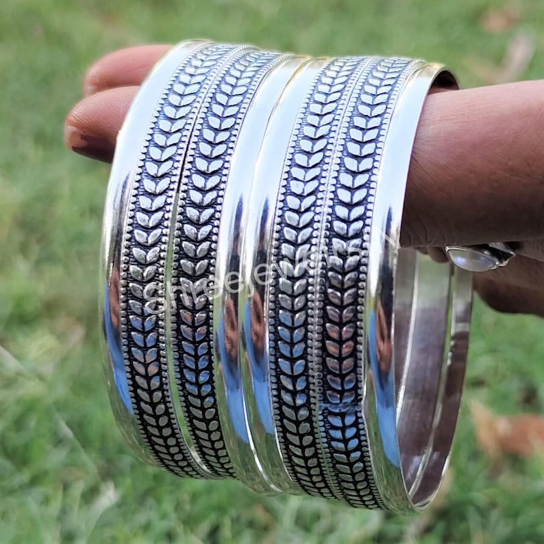 May include: A set of four silver bangles with a detailed leaf pattern. The bangles are thick and shiny, reflecting light. The leaf design is etched into the silver, creating a textured appearance. The bangles are displayed against a green background.