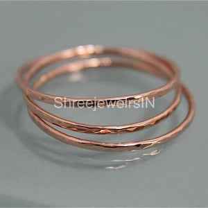 Puede incluir: Tres anillos martillados de oro rosa apilados. Los anillos tienen una superficie texturizada y un diseño simple y elegante. Los anillos se muestran sobre una superficie gris claro.