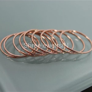 Pode incluir: Uma coleção de anéis finos em ouro rosa dispostos em uma linha ligeiramente curva. Os anéis são de design simples e parecem ser de metal. O fundo é um cinza azulado claro e suave.