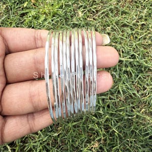Op de afbeelding: Een verzameling zilverkleurige armbanden met een getextureerde, gehamerde afwerking. De armbanden zijn op elkaar gestapeld en reflecteren licht. De sieraden worden tegen een achtergrond van groen gras gehouden.
