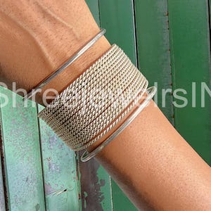 May include: A silver-toned bracelet set. The image shows a stack of woven chain bracelets and two smooth, thin bangles. The bracelets are worn on the wrist against a green background.