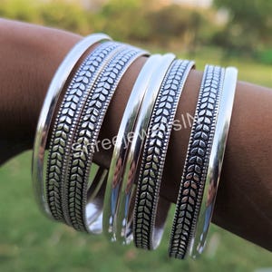 May include: A collection of silver bangles. Some bangles have a smooth, polished finish, while others feature a detailed leaf pattern. The bangles are displayed on a person's arm, showcasing their design and texture. The jewelry is set against a blurred green background.