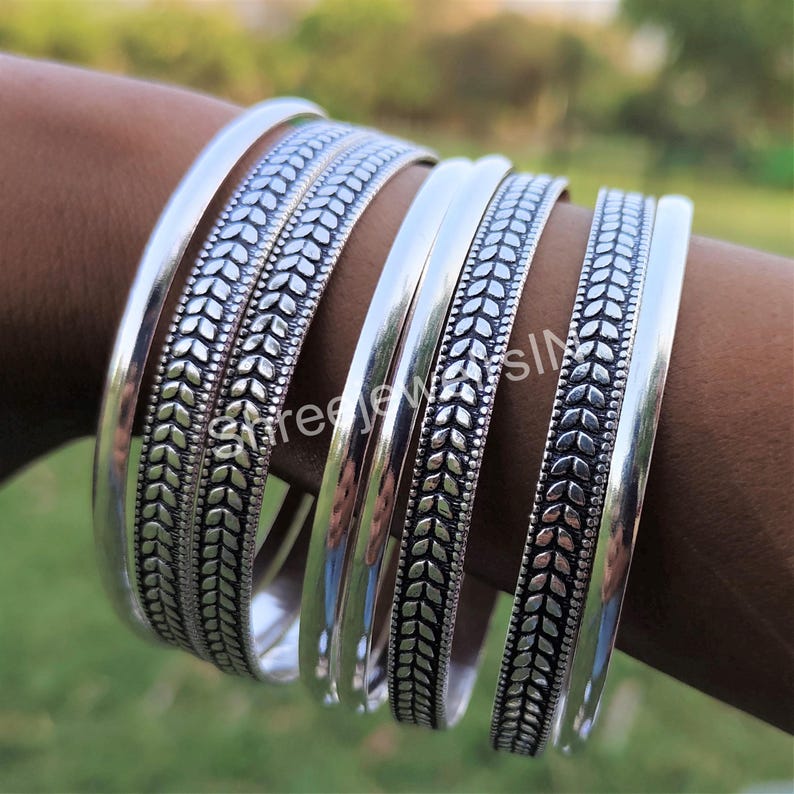 May include: A collection of silver bangles. Some bangles have a smooth, polished finish, while others feature a detailed leaf pattern. The bangles are displayed on a person's arm, showcasing their design and texture. The jewelry is set against a blurred green background.