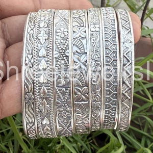 May include: A set of silver bangles featuring detailed floral and geometric designs. The bangles are stacked, highlighting the intricate patterns and textures. The silver colour provides a classic and elegant appearance.