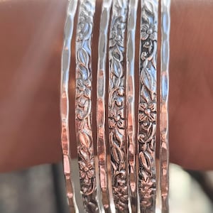 Peut inclure: Ensemble de sept bracelets argent&eacute;s, chacun orn&eacute; d'un motif floral d&eacute;taill&eacute;. Les bracelets sont empil&eacute;s sur un poignet, mettant en valeur le design complexe de fleurs et de feuilles. La lumi&egrave;re se refl&egrave;te sur le m&eacute;tal poli.