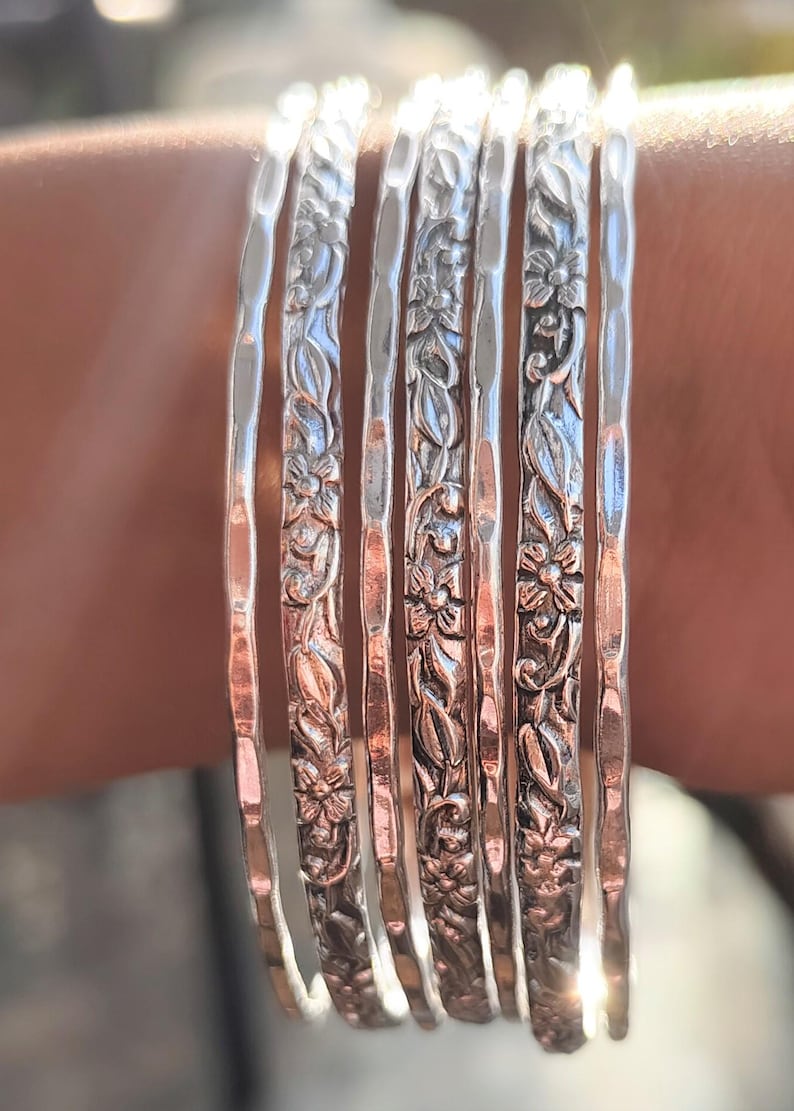 Peut inclure: Ensemble de sept bracelets argent&eacute;s, chacun orn&eacute; d'un motif floral d&eacute;taill&eacute;. Les bracelets sont empil&eacute;s sur un poignet, mettant en valeur le design complexe de fleurs et de feuilles. La lumi&egrave;re se refl&egrave;te sur le m&eacute;tal poli.