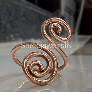 Op de afbeelding: Een koperkleurige ring met een uniek spiraalontwerp. De ring heeft twee prominente spiralen, de ene groter dan de andere, verbonden door een band. Het metaal heeft een gepolijste afwerking, die licht reflecteert. De ring is een sieraad.