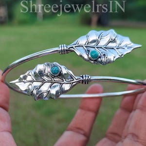 May include: A silver-toned bracelet featuring leaf-shaped designs with turquoise-colored stones. The leaves are detailed with veining and are connected by a curved band. The bracelet is open-ended and appears to be adjustable.