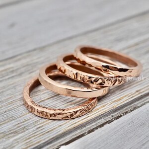 May include: A set of four rose gold rings arranged in a row. Each ring features a distinct surface texture, ranging from a hammered finish to a smooth polished band and a rough, irregular hammered pattern, creating a varied metallic appearance.