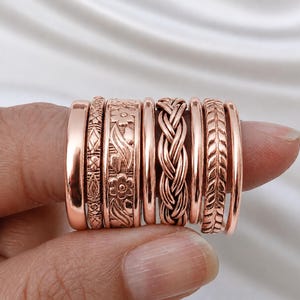 May include: A set of eight rose gold-toned rings featuring various designs, including smooth bands, a geometric pattern, a floral motif, a braided texture, and a leaf-like pattern. The rings are arranged side-by-side, showcasing their metallic finish.