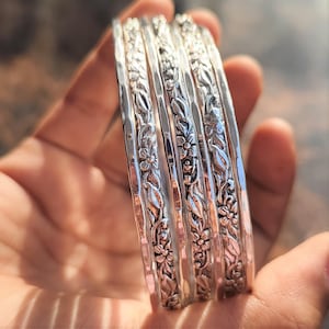 Op de afbeelding: Een set van vijf zilveren armbanden met bloemen- en bladerpatronen. De armbanden worden in een hand gehouden, waarbij de gedetailleerde ontwerpen zichtbaar zijn. Ze hebben een gehamerde textuur en een glanzende afwerking.