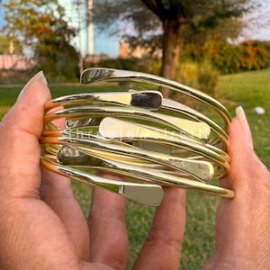 May include: A stack of gold-colored metal arm cuffs. The cuffs have a smooth, polished finish and a curved design. The bracelets are held in a person's hand against a blurred green background.