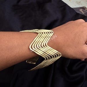 Op de afbeelding: Goudkleurige metalen manchetarmband met een zigzagontwerp. De armband wordt om een arm gedragen en benadrukt de geometrische vorm en de gepolijste afwerking. De manchet is een statement-stuk en voegt een vleugje moderne stijl toe.