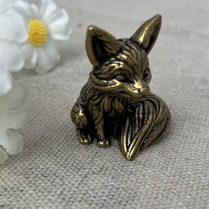 May include: A small, detailed brass fox figurine with a curled tail. The fox is posed in a sitting position, with intricate fur detailing. White daisy flowers with yellow centers are in the background.