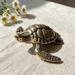 May include: A small, metallic, gold-toned sea turtle figurine with a detailed, segmented shell and textured flippers. The turtle is depicted in a swimming pose, showcasing a weathered, antique-style finish with dark highlights in the crevices.