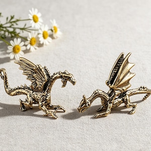 May include: Two small, metallic gold-toned dragon figurines with detailed scales, textured wings, and long tails. The dragons are posed in a standing position with their heads turned, showcasing intricate craftsmanship and a polished finish.
