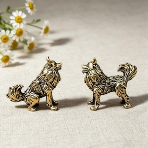 May include: Two small, detailed brass figurines of dogs with textured fur and harness-like markings. One dog is depicted in a seated position with its tail curled, while the other stands with its head turned and tail raised, both showing a metallic finish.