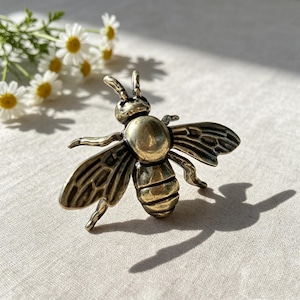 May include: A metallic bee-shaped object with a brass-toned finish, featuring detailed wings with etched lines, a rounded thorax, and a segmented abdomen. The bee is positioned with its wings spread, showing a dark patina in the recessed areas.