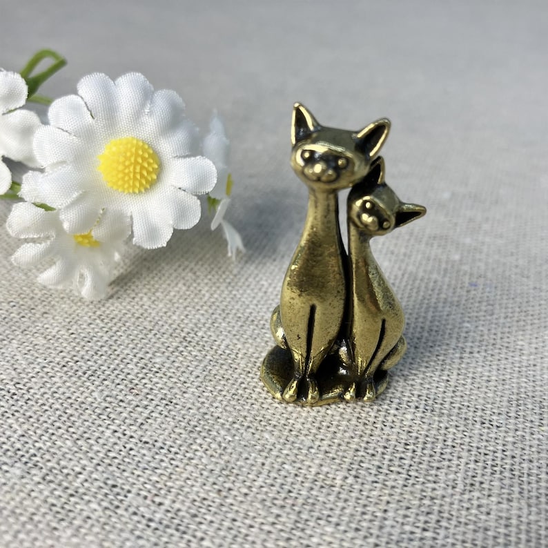 Op de afbeelding: Een bronzen kattenbeeldje dat twee katten voorstelt die dicht bij elkaar zitten. De katten hebben puntige oren en staan op een getextureerd, lichtgrijs oppervlak. Witte madeliefjes met gele centra zijn op de achtergrond.