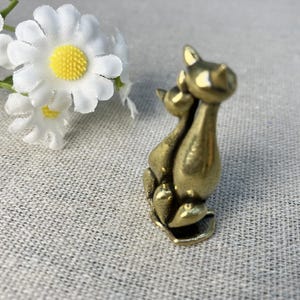 Op de afbeelding: Een kleine, bronskleurige kattenfiguur die twee dicht bij elkaar zittende katten afbeeldt. De katten hebben puntoren en zitten op een kleine basis. Witte madeliefjes met gele centra zijn op de achtergrond.