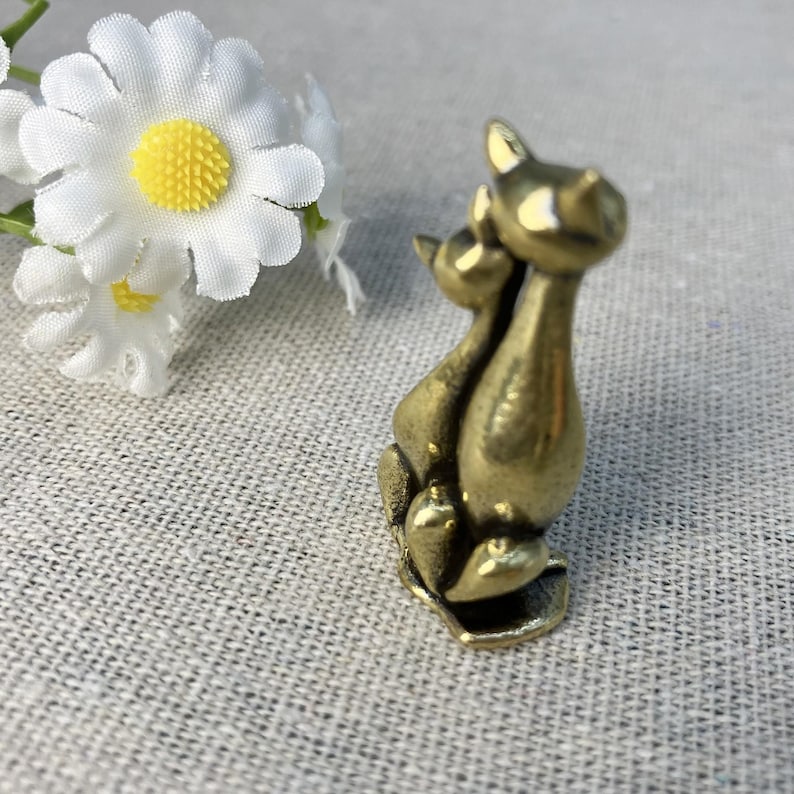 Op de afbeelding: Een kleine, bronskleurige kattenfiguur die twee dicht bij elkaar zittende katten afbeeldt. De katten hebben puntoren en zitten op een kleine basis. Witte madeliefjes met gele centra zijn op de achtergrond.
