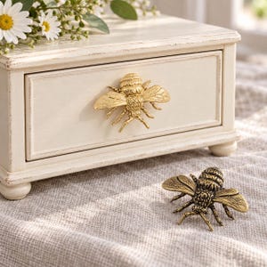 May include: A cream-colored wooden box with a single drawer featuring a gold-toned bee-shaped handle. A matching gold-toned bee-shaped handle is placed separately on the surface in front of the box. The box has a distressed finish.