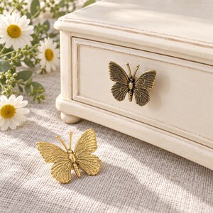 May include: Two butterfly-shaped metal drawer knobs with detailed wing textures. One knob is finished in an antique bronze tone and is attached to a white wooden drawer, while the other is a bright gold tone resting on a textured surface.