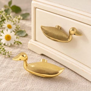 May include: Two brass-toned metal drawer knobs shaped like ducks. One knob is attached to a white wooden drawer, while the other rests on a textured surface. The duck-shaped hardware features a smooth, metallic finish and a distinct head and body.