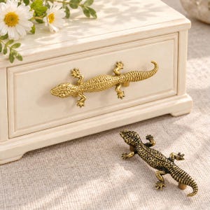 May include: A cream-colored wooden drawer features a gold-toned lizard-shaped metal handle. A second, darker bronze-toned lizard-shaped handle is positioned in front of the drawer. White daisy flowers are visible at the top left corner.