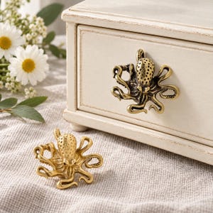 May include: Two brass-colored metal cabinet knobs shaped like octopuses with textured heads and curved tentacles. One knob is attached to the front of a cream-colored wooden drawer, while the other sits on a textured fabric surface.