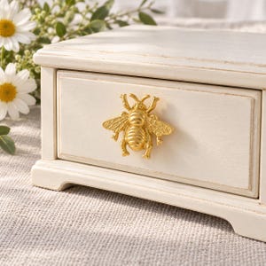 May include: A small, rectangular, off-white wooden storage box featuring a single drawer. The drawer is equipped with a prominent, three-dimensional gold-colored metal bee-shaped handle centered on its front panel.